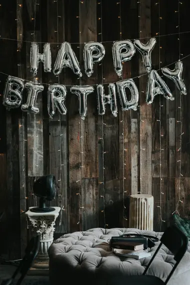 Silberne Happy-Birthday-Ballons vor einer Holzwand mit Lichterketten